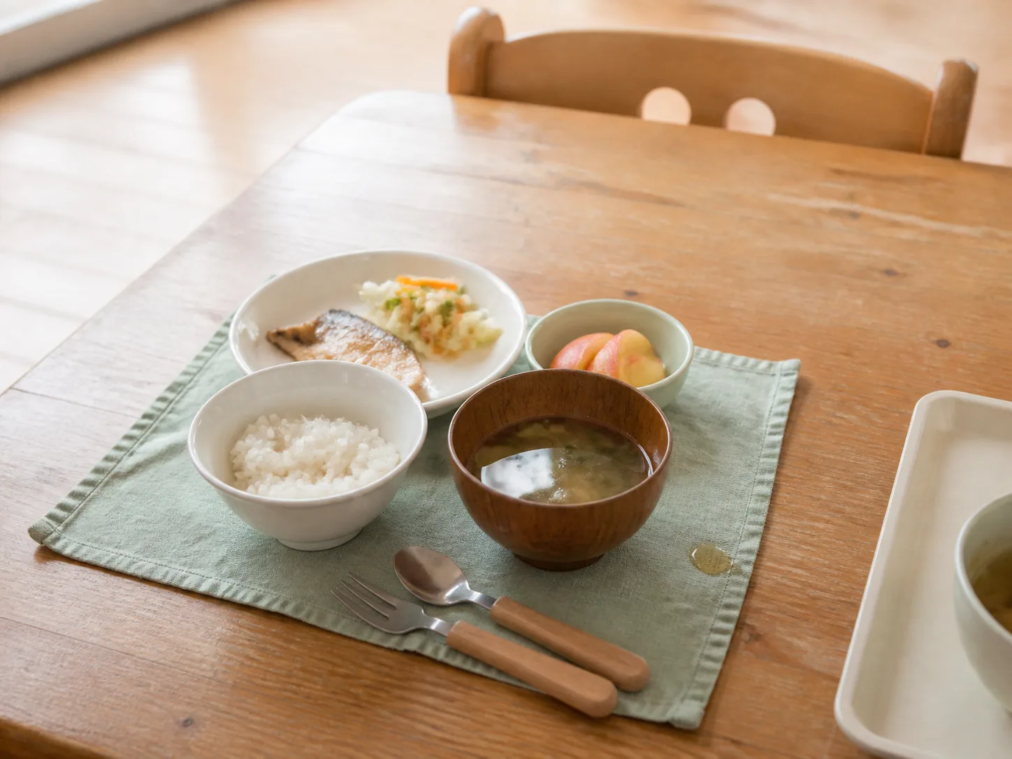 木のテーブルに配膳された給食。ごはん、汁物、焼き魚、副菜、果物