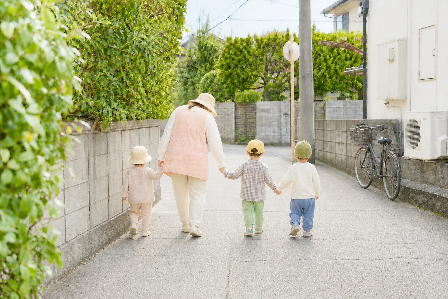 住宅街の道を、保育士と手をつないで歩く子どもたちの後ろ姿